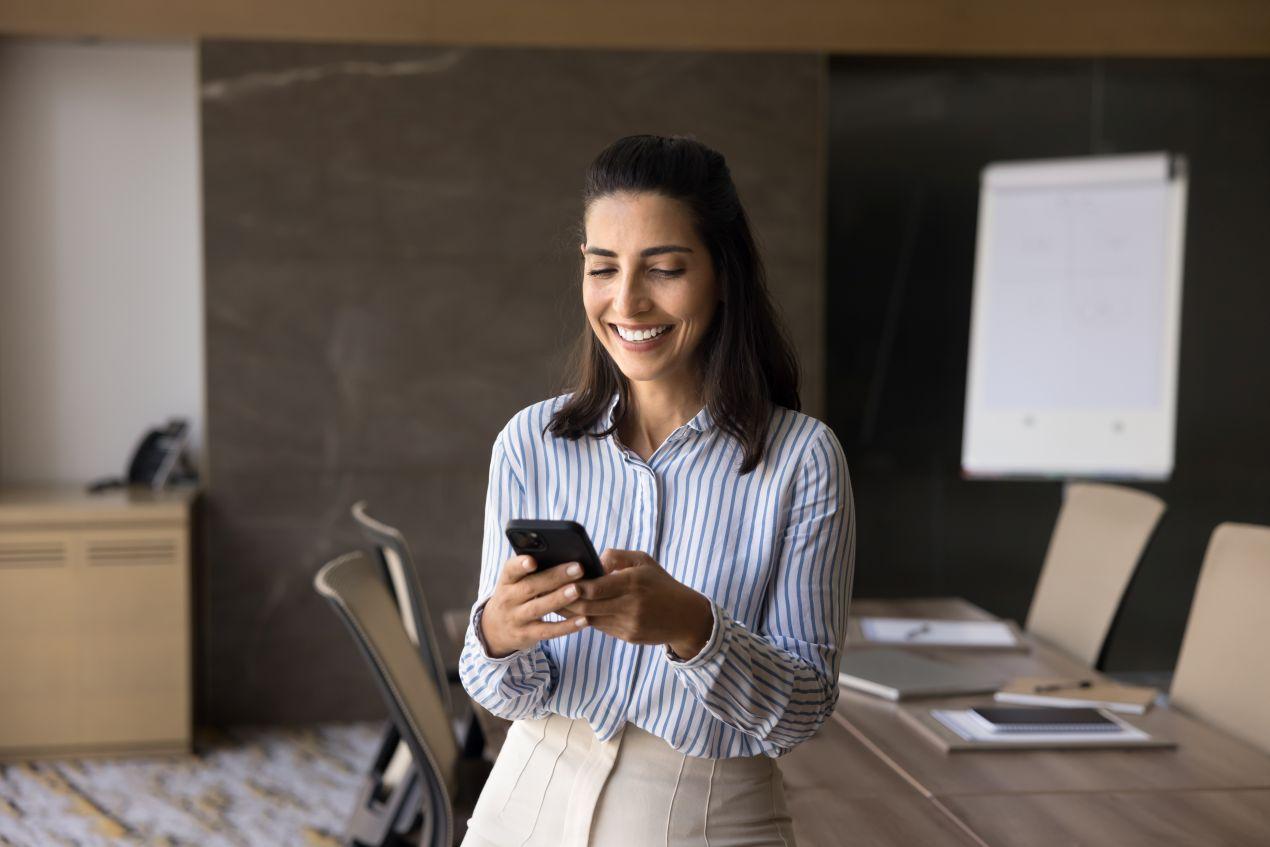 Lächelnde Frau nutzt E-Learning Content auf ihrem Smartphone in einem modernen Büro. Mobiles Lernen mit einem Learning Management System (LMS).